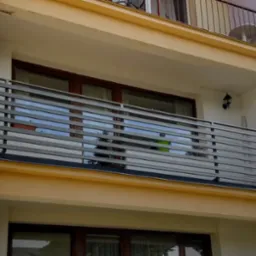 Szara, pozioma balustrada balkonowa z metalu, z widocznym fragmentem elewacji budynku w ciepłej kolorystyce. Balkon na drugim piętrze z oknami.