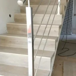 Schody z drewnianymi stopniami i nowoczesną balustradą ze stali nierdzewnej z drewnianą poręczą. Widoczne elementy montażowe i fragment podłogi w trakcie wykończenia.