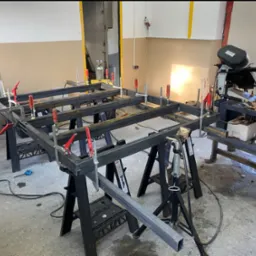 Metalowa konstrukcja spawana, złączona ściskami stolarskimi, na warsztacie. W tle maszyna do cięcia metalu i butla z gazem.