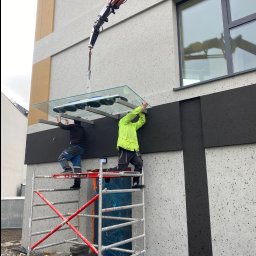 GLOBALSKILL SPÓŁKA Z OGRANICZONĄ ODPOWIEDZIALNOŚCIĄ - Montaż szklanego daszku nad wejściem do budynku przez dwóch pracowników na rusztowaniu, z użyciem dźwigu z przyssawkami. Fasada budynku w odcieniach szarości.