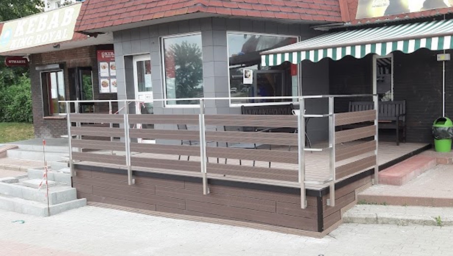 Zewnętrzny taras z balustradą z poziomych desek w kolorze brązowym i metalowymi słupkami, przy lokalu gastronomicznym z zielonym daszkiem.
