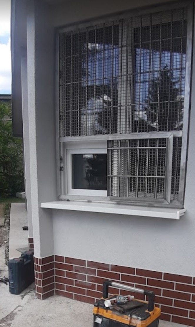 Metalowa krata okienna z uchylną częścią, zamontowana na zewnątrz budynku z ceglaną podmurówką. Widoczne narzędzia w skrzynce.