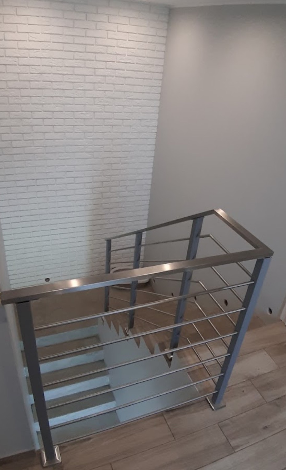 Metalowa balustrada schodowa z poziomymi prętami, widok z góry, biała ściana z cegły w tle, drewnopodobne płytki na podłodze, nowoczesny design.