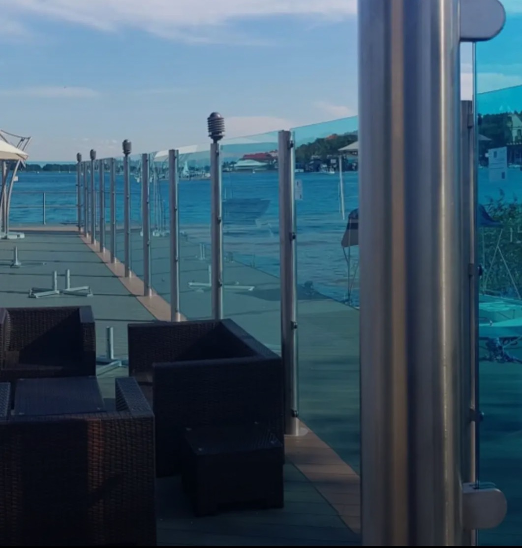 Szklane balustrady z metalowymi słupkami na tarasie z widokiem na wodę. W tle widać budynki. Elementy mebli ogrodowych na pierwszym planie.