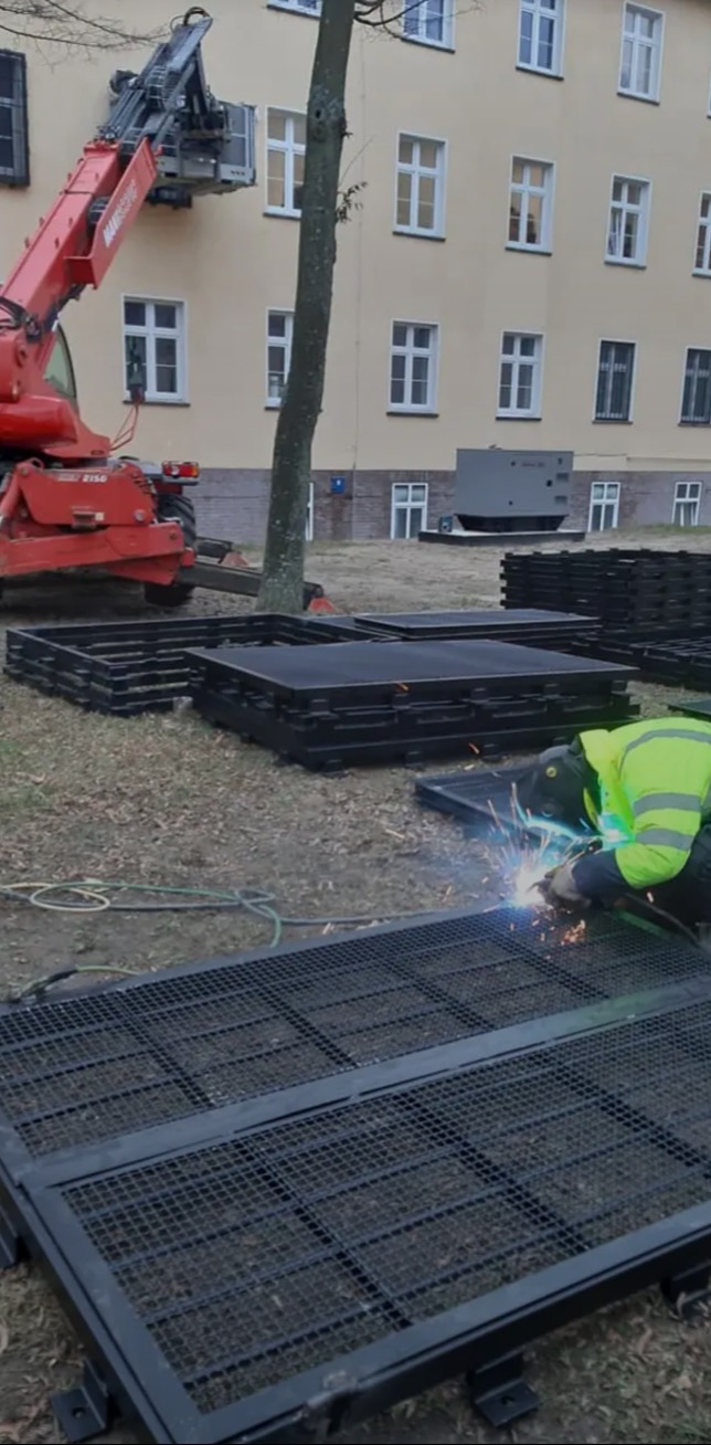 Pracownik w odblaskowej kamizelce spawa metalową kratę na trawie. W tle budynek i podnośnik teleskopowy.