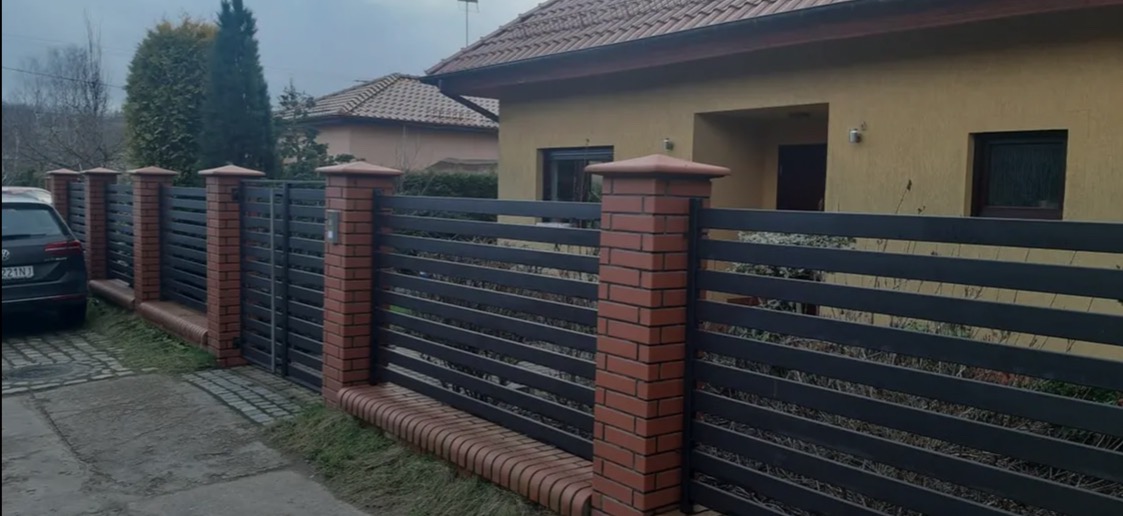 Nowoczesne ogrodzenie metalowe z elementami ceglanymi, poziome przęsła, widok z zewnątrz na tle domu, zieleń w tle, pochmurny dzień.