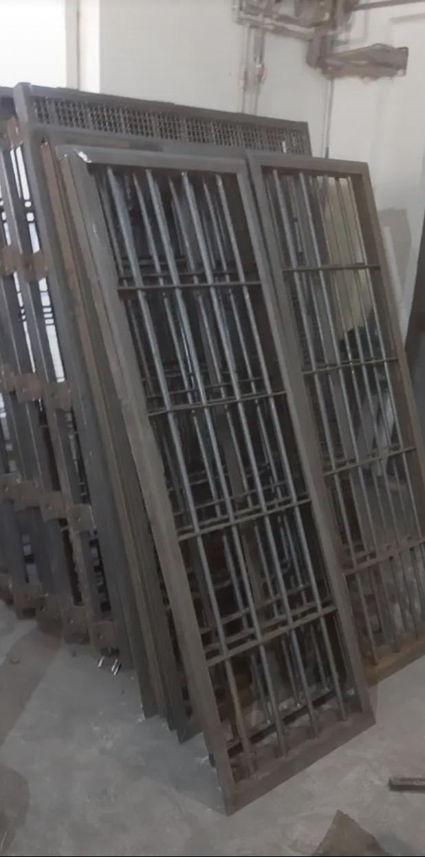 Stos metalowych ram z kratami, prawdopodobnie elementów ogrodzenia lub balustrady, w szarym odcieniu, opartych o ścianę w warsztacie.