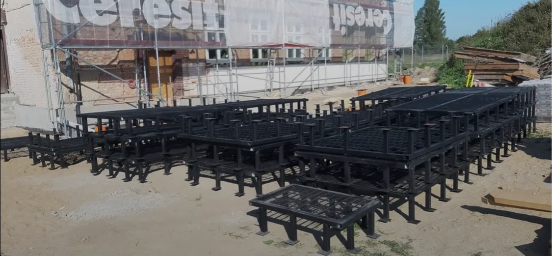 Szkielety metalowe pod konstrukcje, czarne, ustawione w stosy na placu budowy. Widoczne rusztowania i budynek w tle. Elementy przygotowane do montażu.