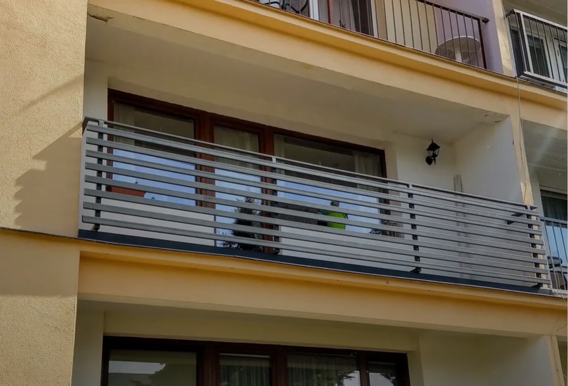 Szara, pozioma balustrada balkonowa z metalu, z widocznym fragmentem elewacji budynku w ciepłej kolorystyce. Balkon na drugim piętrze z oknami.