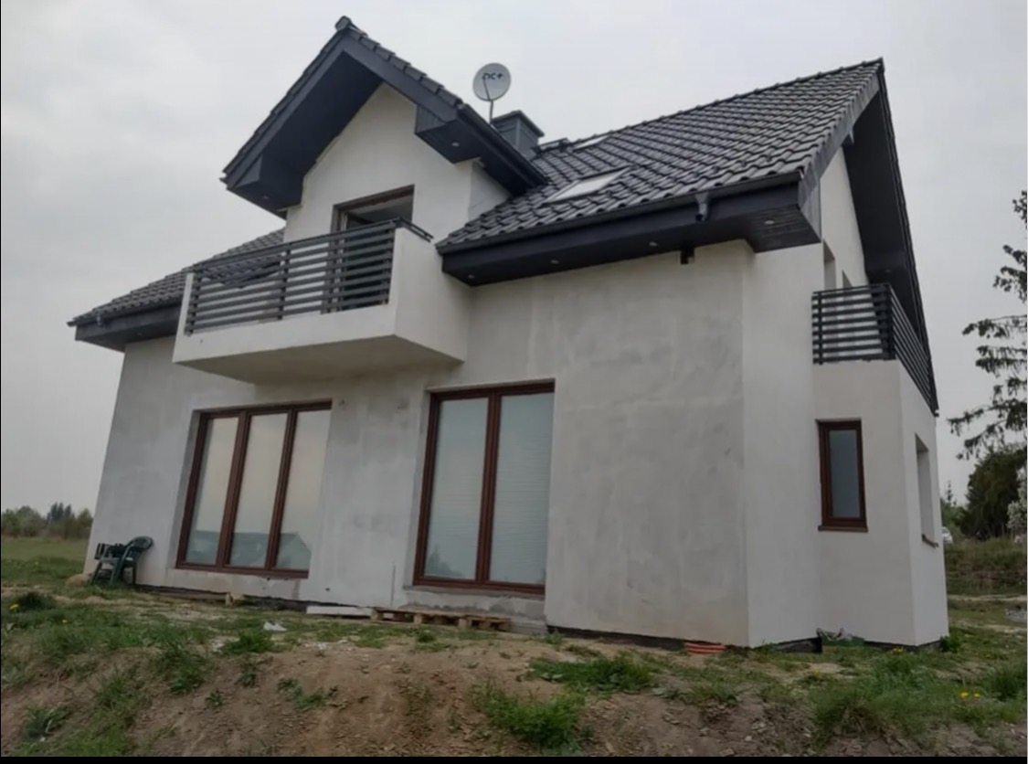 Dwukondygnacyjny dom z balkonem i szarą balustradą, widoczny z zewnątrz. Elewacja w odcieniach bieli, ciemna dachówka. Uwagę zwraca satelita na dachu i krzesło na trawie.