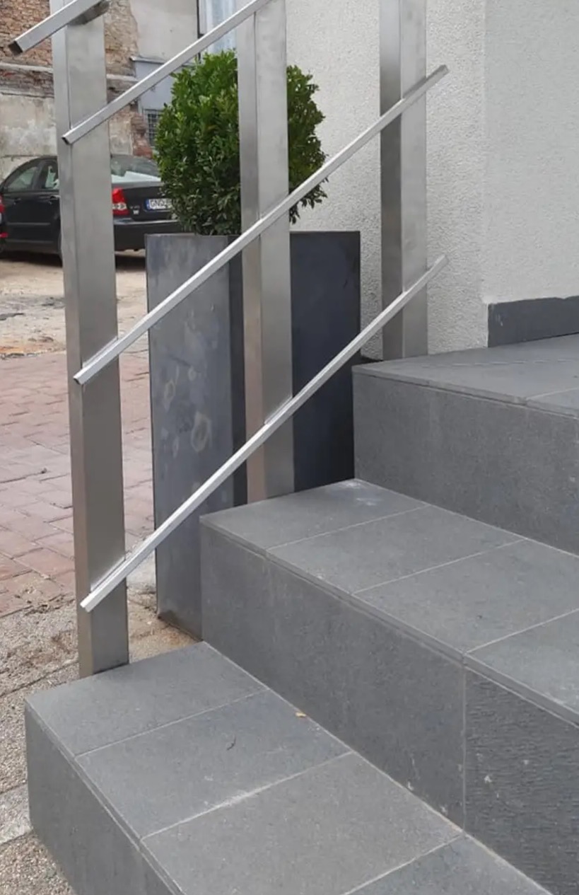 Nowoczesne, metalowe poręcze schodowe z subtelnymi, poziomymi elementami, prowadzące do wejścia budynku. Obok schodów widoczna donica z zieloną rośliną.