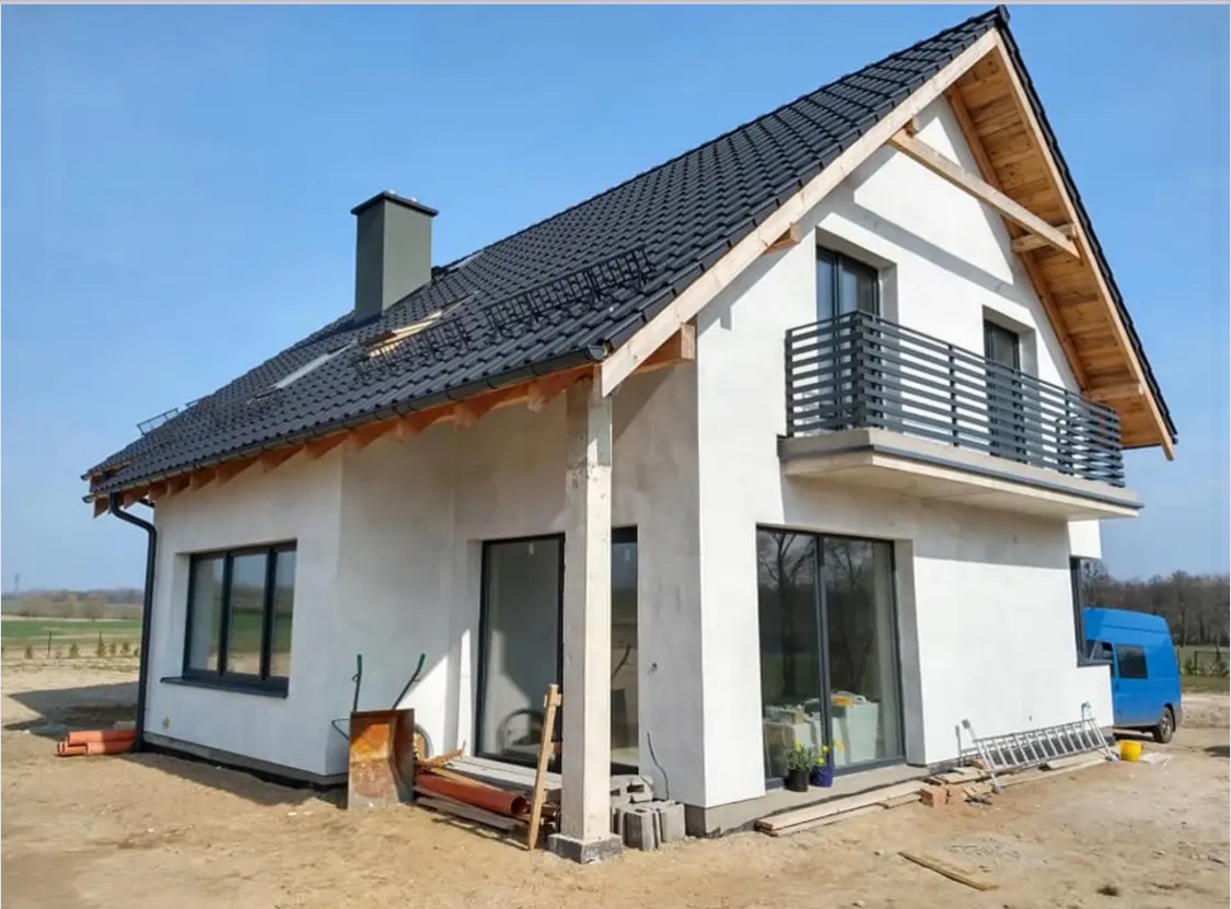 Dom jednorodzinny z czarną balustradą balkonową i dachem z dachówki, w trakcie wykończenia elewacji. Widoczny fragment niebieskiego samochodu dostawczego.