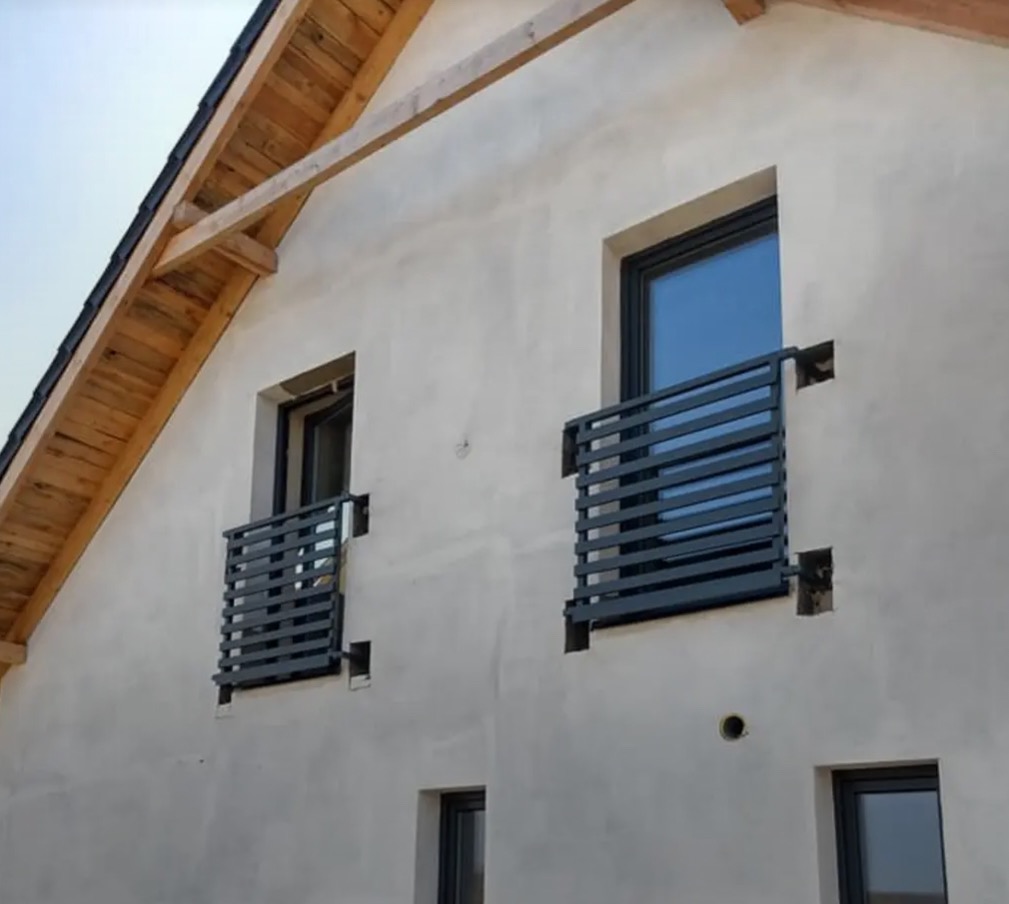 Szare, metalowe balustrady okienne zamontowane na elewacji domu w trakcie budowy. Widoczne surowe drewno konstrukcji dachu i fragmenty niezakończonych prac murarskich.