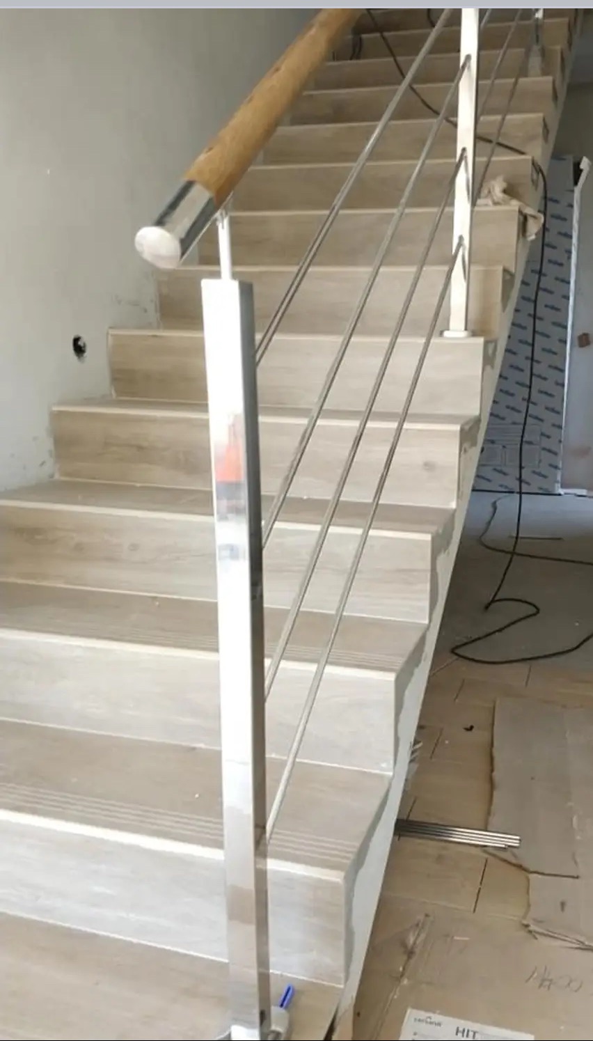 Schody z drewnianymi stopniami i nowoczesną balustradą ze stali nierdzewnej z drewnianą poręczą. Widoczne elementy montażowe i fragment podłogi w trakcie wykończenia.