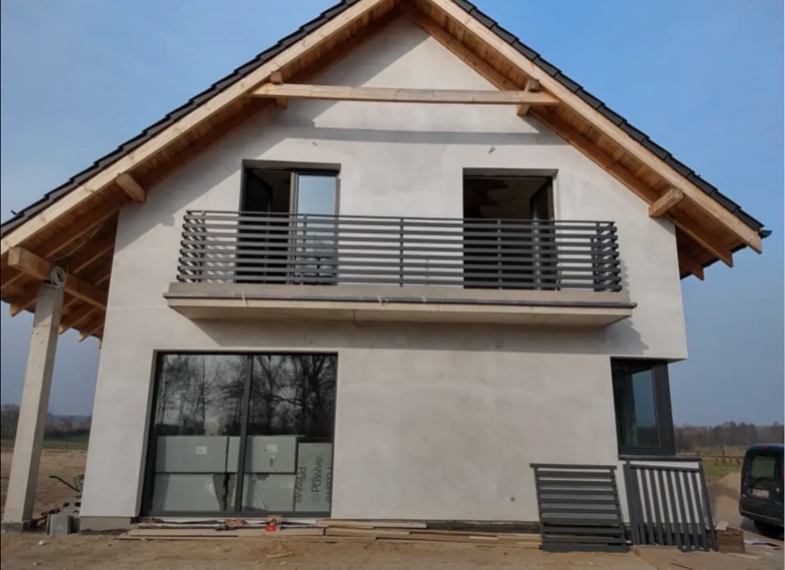 Dom w trakcie budowy z zamontowaną metalową balustradą balkonową. Na dole leżą elementy balustrady. Widoczne surowe wykończenie elewacji.
