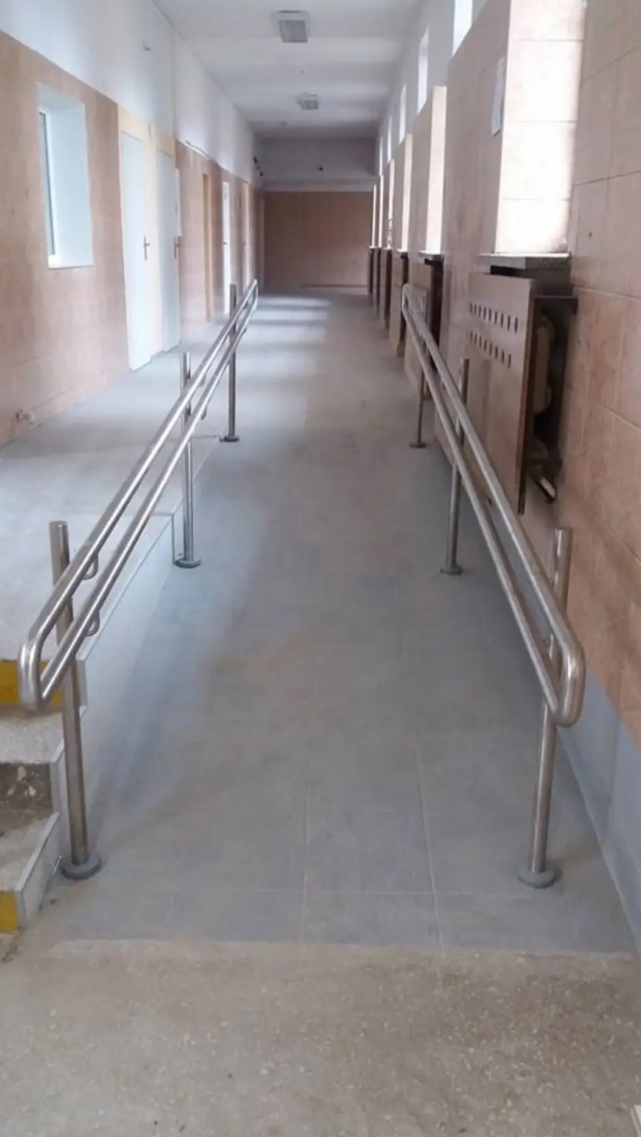 Długi korytarz z rampą i poręczami ze stali nierdzewnej, umożliwiający dostęp dla osób z niepełnosprawnościami. Widoczne płytki na podłodze i ściany w odcieniach beżu.