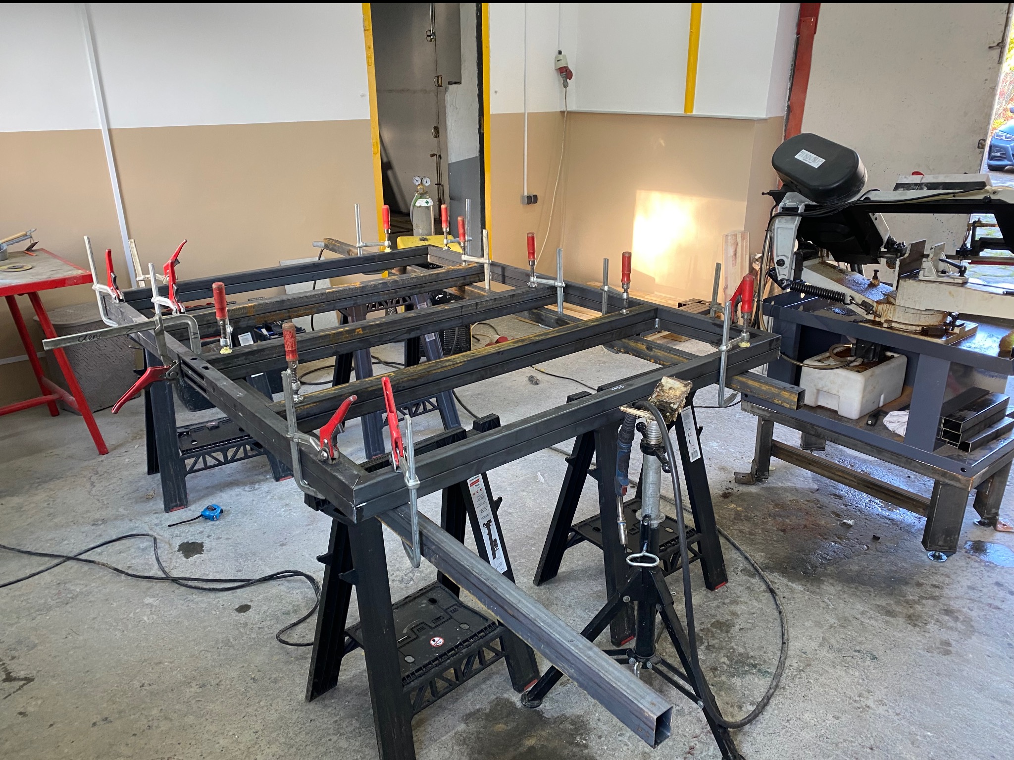 Metalowa konstrukcja spawana, złączona ściskami stolarskimi, na warsztacie. W tle maszyna do cięcia metalu i butla z gazem.