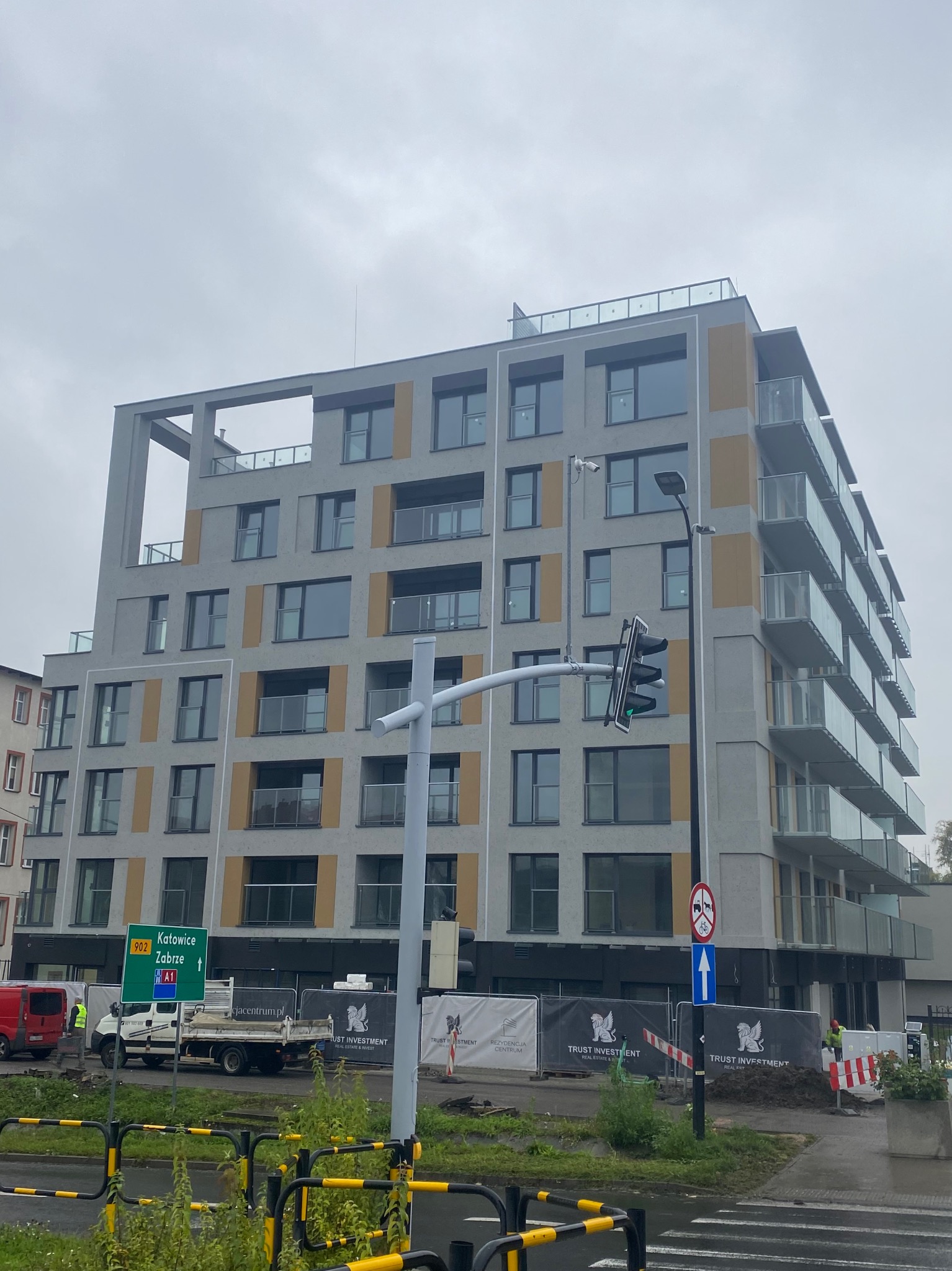 Nowoczesny budynek mieszkalny z balkonami o szklanych balustradach, widziany z ulicy w pochmurny dzień. W tle sygnalizacja świetlna i znaki drogowe.