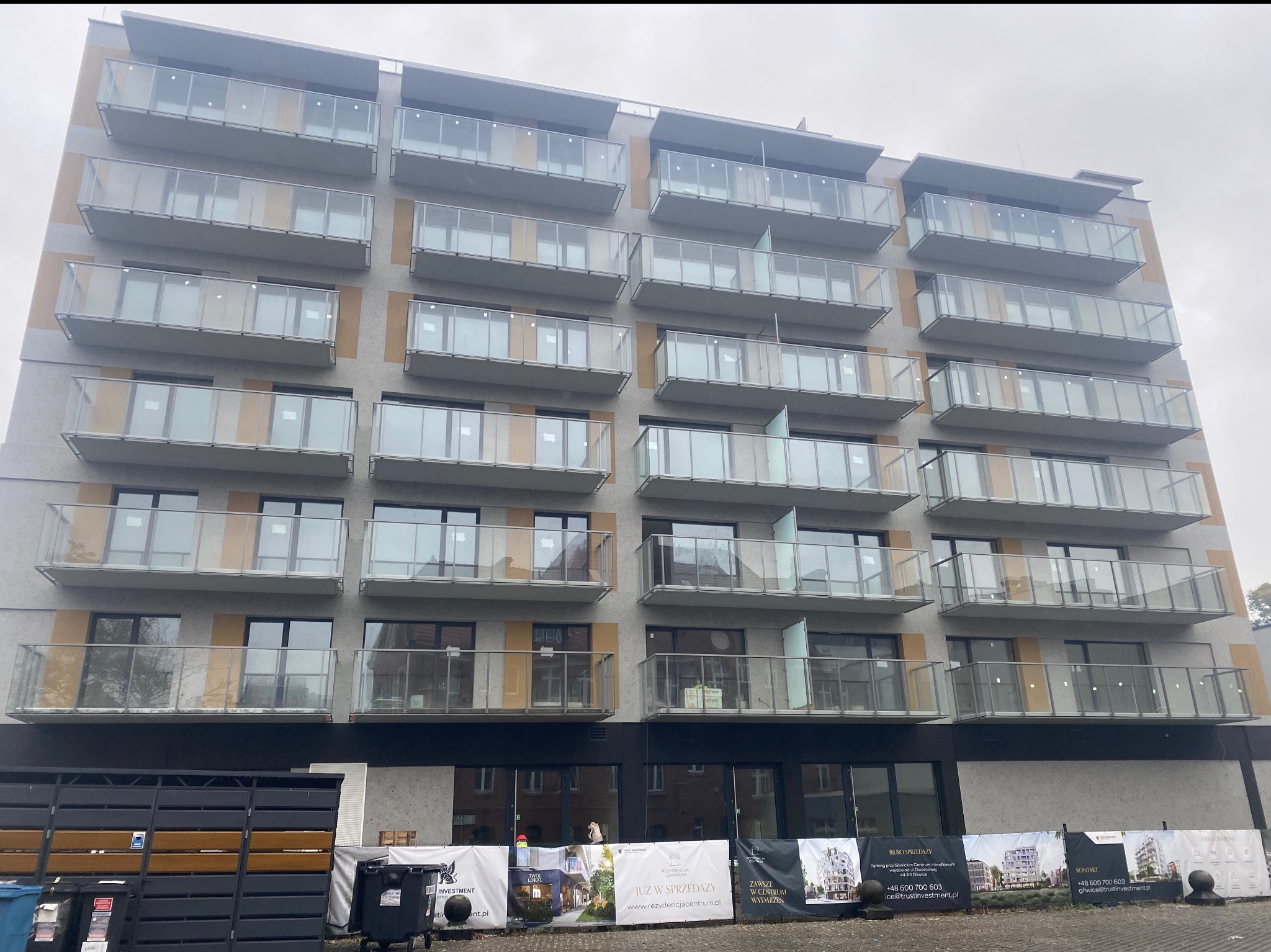 Wielopiętrowy budynek z balkonami z balustradami szklanymi, widok z dołu na elewację z szarymi panelami i żółtymi akcentami. Na dole banery reklamowe.
