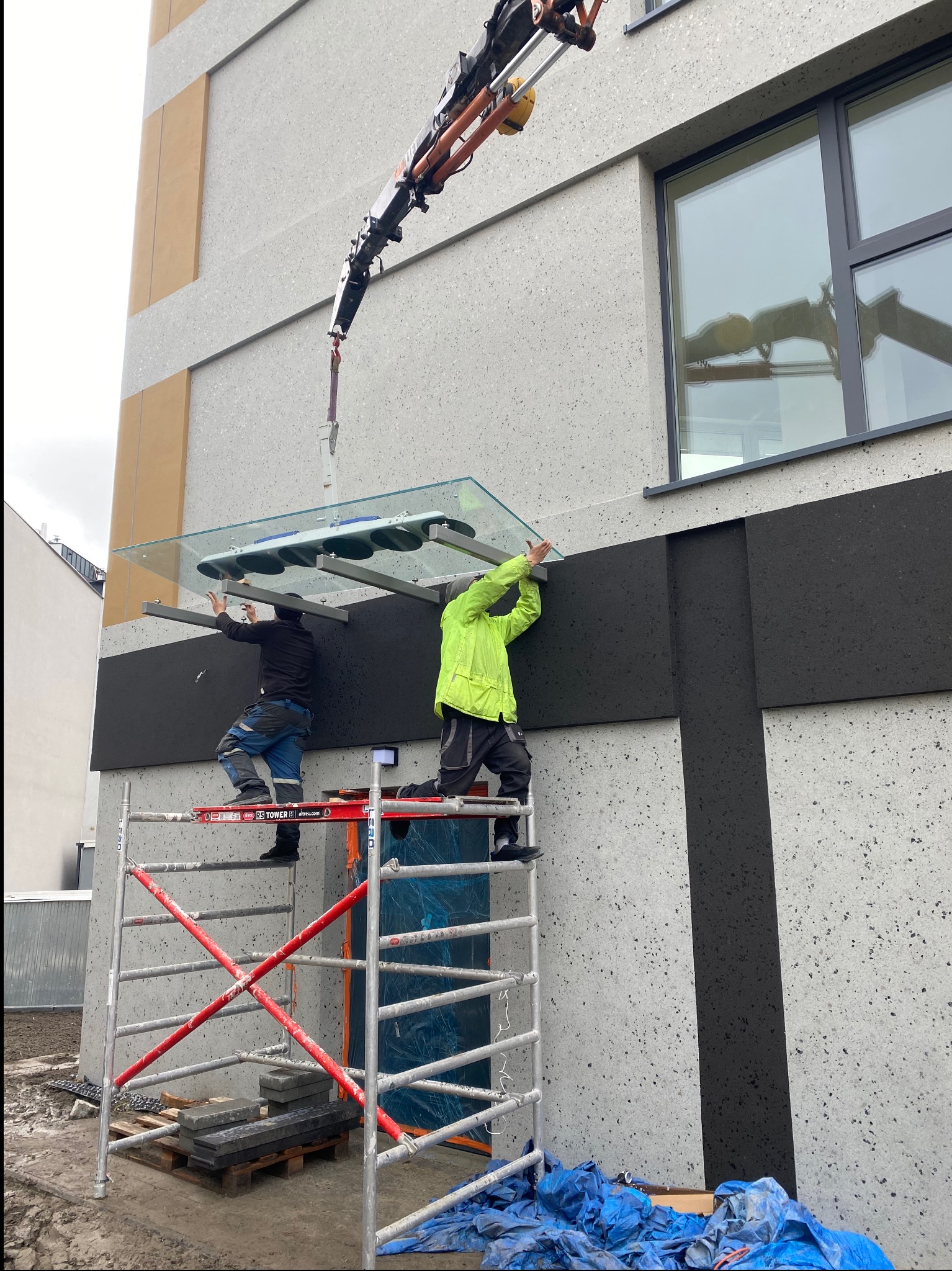 Montaż szklanego daszku nad wejściem do budynku przez dwóch pracowników na rusztowaniu, z użyciem dźwigu z przyssawkami. Fasada budynku w odcieniach szarości.