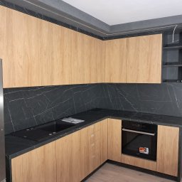 ZIBI Zbigniew Bizoń - Nowoczesna kuchnia w trakcie montażu: jasne drewniane fronty szafek, czarny blat z marmurkowym wzorem, widoczne elementy instalacji elektrycznej i niedokończone ściany.