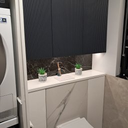 ZIBI Zbigniew Bizoń - Nowoczesna łazienka z zabudową nad toaletą: grafitowe szafki z ryflowanymi frontami, marmurowy backsplash, biały blat z dekoracjami (rośliny, dyfuzor zapachowy), pralka i suszarka w kolumnie obok.