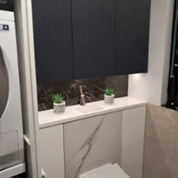 Nowoczesna łazienka z zabudową nad toaletą: grafitowe szafki z ryflowanymi frontami, marmurowy backsplash, biały blat z dekoracjami (rośliny, dyfuzor zapachowy), pralka i suszarka w kolumnie obok.