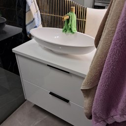 ZIBI Zbigniew Bizoń - Biała szafka łazienkowa z owalną umywalką nablatową, czarne uchwyty, złota bateria i dekoracyjne lustro z metalowymi prętami, widoczne ręczniki.