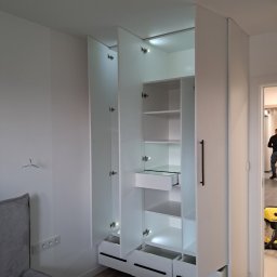 ZIBI Zbigniew Bizoń - Biała szafa wnękowa z oświetleniem LED, otwarte drzwi ukazują półki i szuflady, widoczny fragment łóżka i osoba w tle.