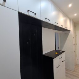 ZIBI Zbigniew Bizoń - Biała zabudowa meblowa z czarnymi akcentami, obejmująca szafki i wieszak na ubrania w przedpokoju, z minimalistycznymi uchwytami i oświetleniem punktowym.