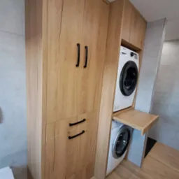 Drewniana zabudowa z szafkami i szufladami, integrująca pralkę i suszarkę w łazience, z wysuwanym blatem roboczym między urządzeniami.