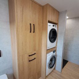 ZIBI Zbigniew Bizoń - Zabudowa w łazience z drewna z pralką i suszarką, czarne uchwyty, jasna podłoga, widoczny fragment toalety i umywalki.