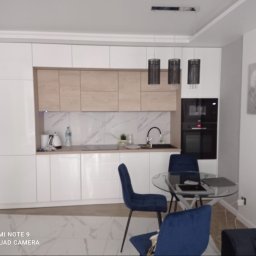 ZIBI Zbigniew Bizoń - Nowoczesna kuchnia z białymi szafkami i drewnianymi akcentami, marmurowy backsplash, okrągły stół ze szklanym blatem i niebieskimi krzesłami, minimalistyczne lampy wiszące.