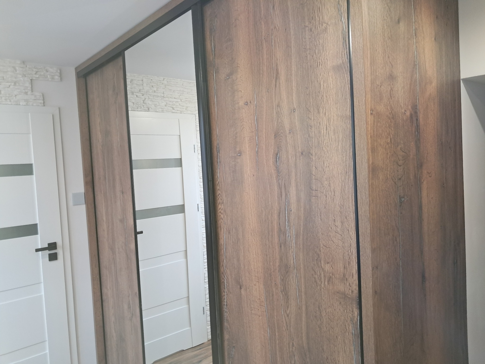 Szafa wnękowa z przesuwnymi drzwiami z lustrem i okleiną w kolorze ciemnego drewna, widoczna na tle białych drzwi i ściany z dekoracyjną cegłą.