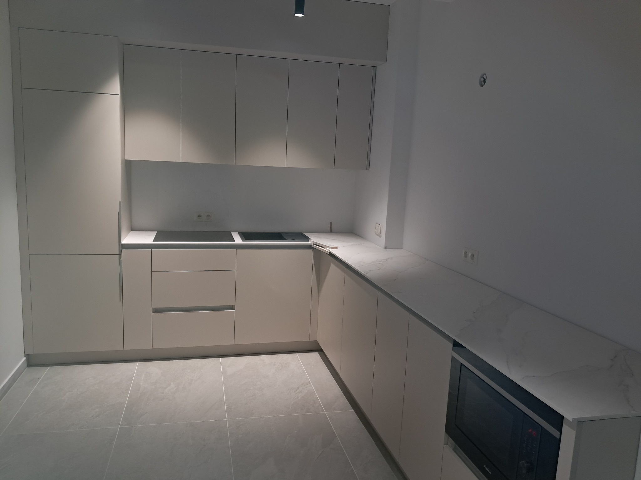 Jasna, minimalistyczna kuchnia z zabudową sięgającą sufitu, blatem imitującym marmur i płytą indukcyjną, oświetlona punktowym światłem.