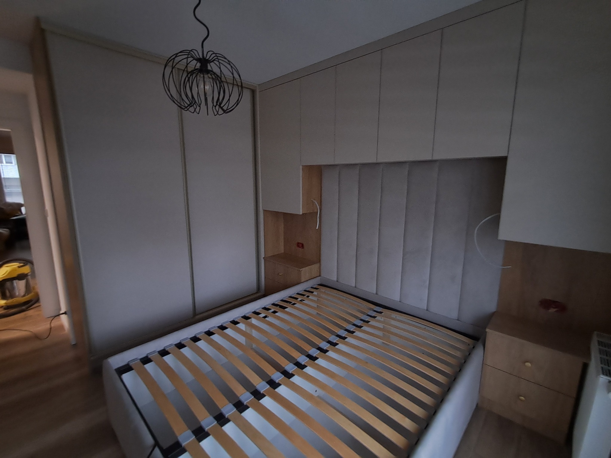 Komplet mebli do sypialni: szafa przesuwna z jasnymi frontami, łóżko z tapicerowanym wezgłowiem i drewnianym stelażem oraz szafki nocne z szufladami, oświetlone dekoracyjną lampą wiszącą.