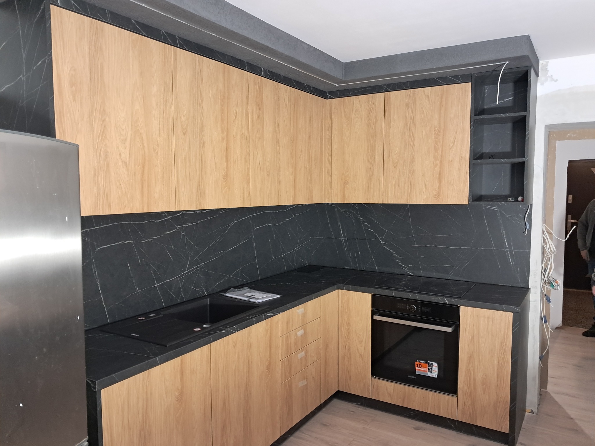 Nowoczesna kuchnia w trakcie montażu: jasne drewniane fronty szafek, czarny blat z marmurkowym wzorem, widoczne elementy instalacji elektrycznej i niedokończone ściany.
