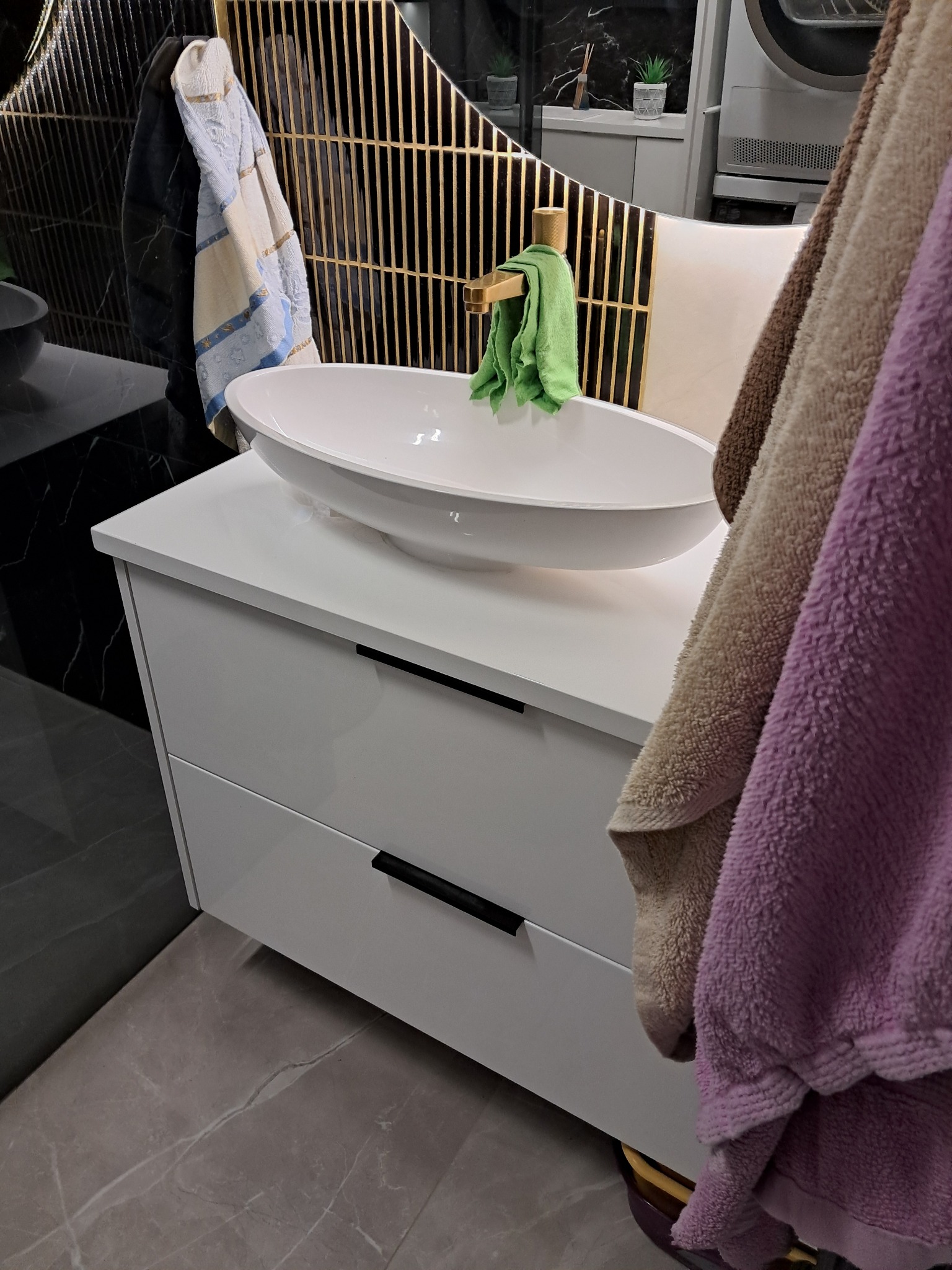 Biała szafka łazienkowa z owalną umywalką nablatową, czarne uchwyty, złota bateria i dekoracyjne lustro z metalowymi prętami, widoczne ręczniki.