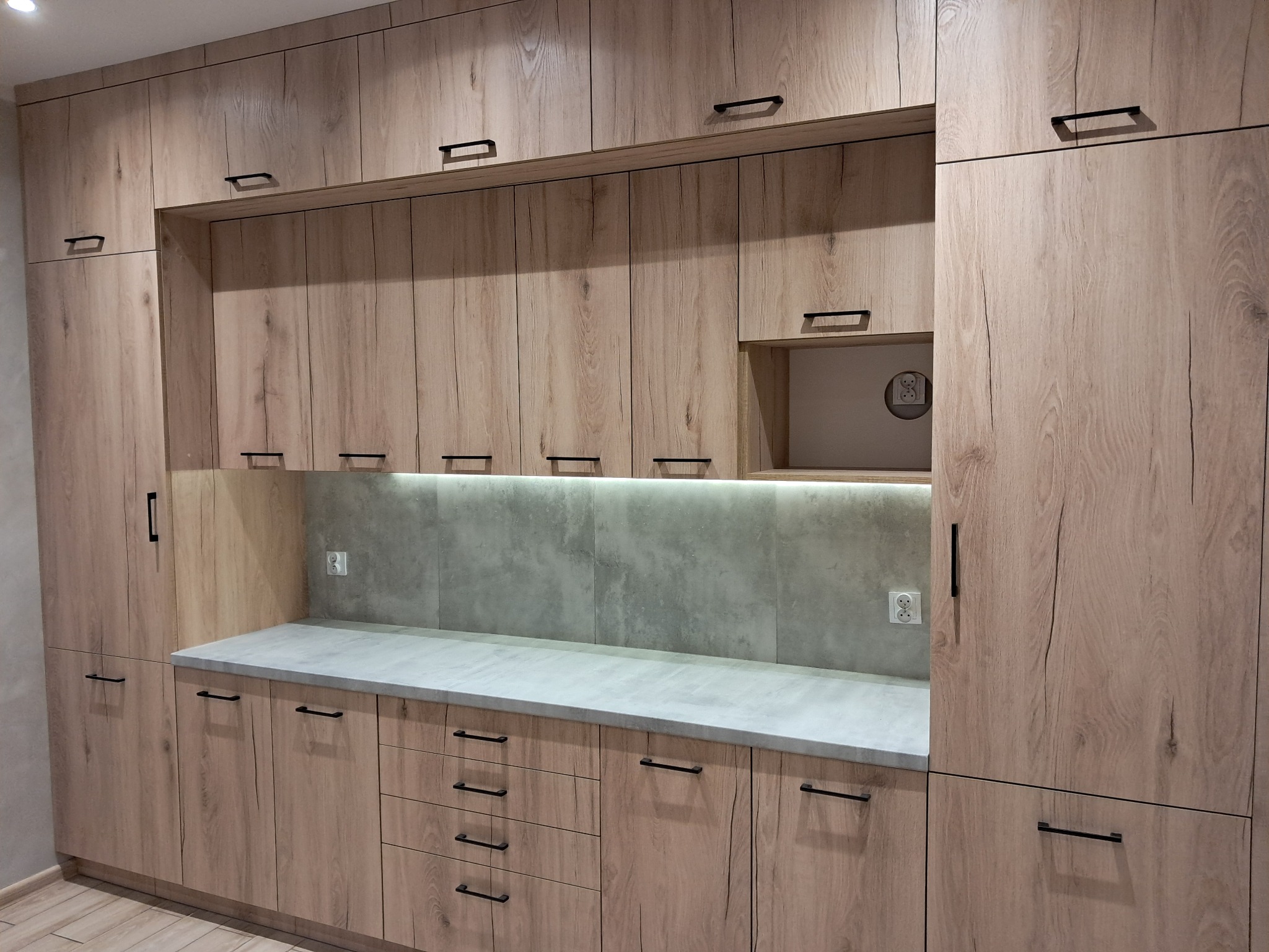 Jasna zabudowa kuchenna z drewnopodobnymi frontami, czarnymi uchwytami i szarym blatem, oświetlenie LED pod szafkami wiszącymi, gniazdko elektryczne na ścianie.