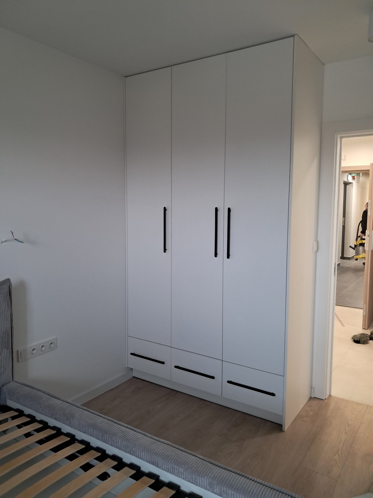 Biała szafa wnękowa z trzema parami drzwi i trzema szufladami z czarnymi uchwytami, dopasowana do białych ścian w sypialni, widok z perspektywy łóżka z szarym zagłówkiem.