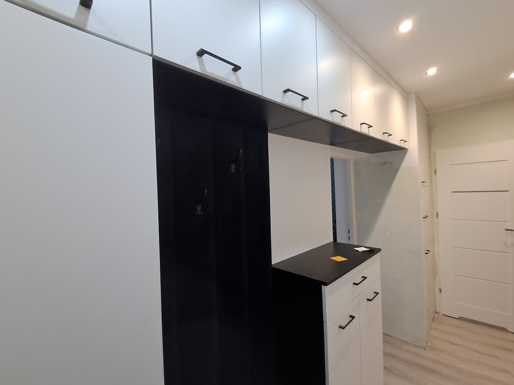 Biała zabudowa meblowa z czarnymi akcentami, obejmująca szafki i wieszak na ubrania w przedpokoju, z minimalistycznymi uchwytami i oświetleniem punktowym.
