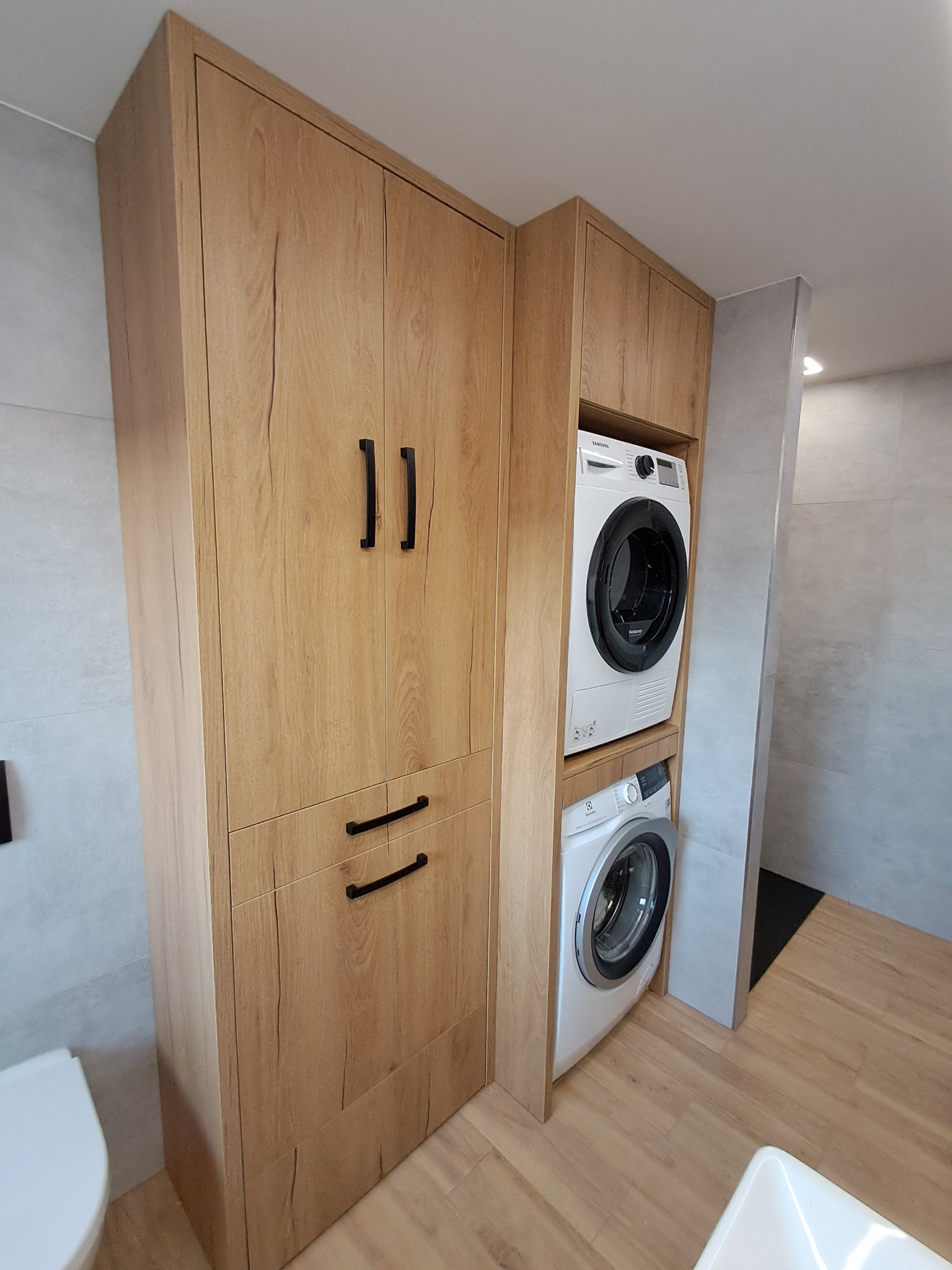 Zabudowa w łazience z drewna z pralką i suszarką, czarne uchwyty, jasna podłoga, widoczny fragment toalety i umywalki.