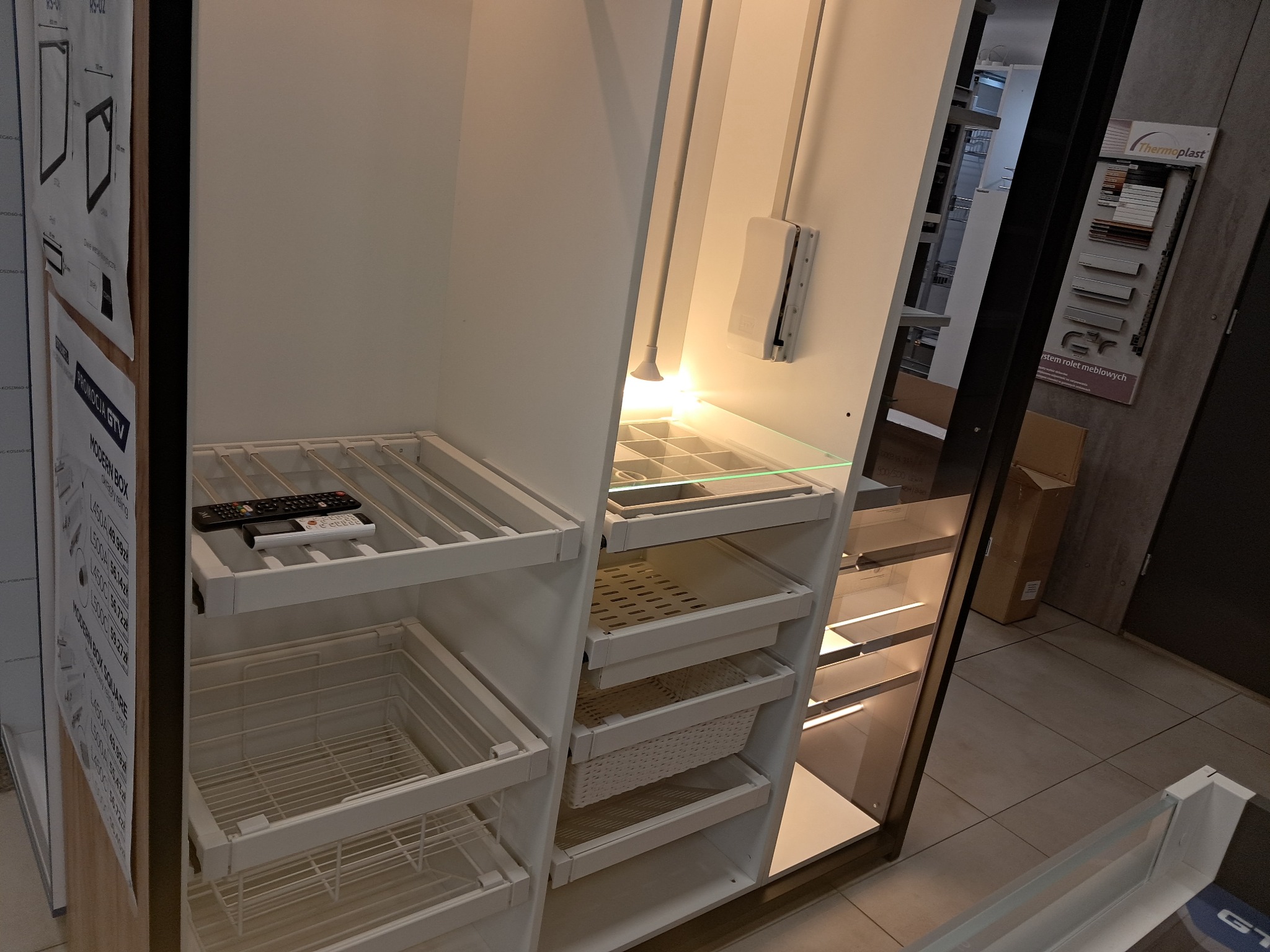Wnętrze szafy wnękowej z wysuwanymi koszami, półkami i oświetleniem, widoczne dwa piloty na półce, ekspozycja w salonie meblowym z katalogami producentów w tle.
