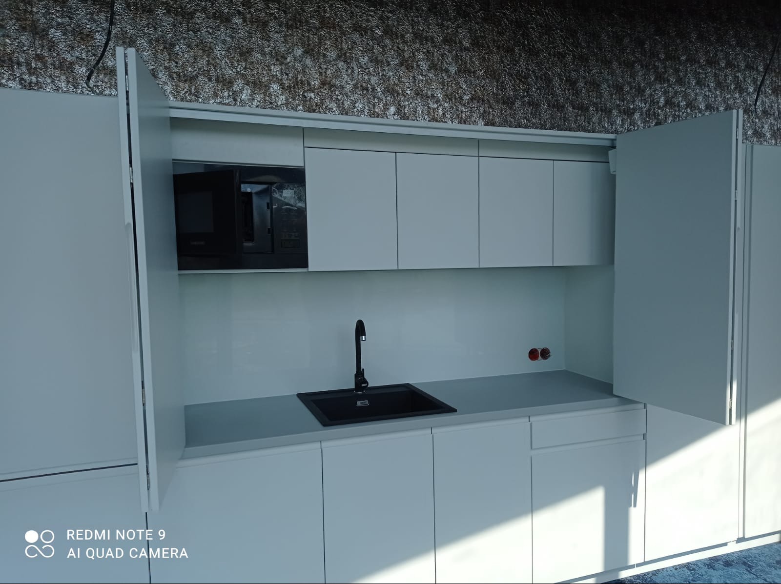 Minimalistyczna, biała kuchnia na wymiar z czarnym zlewem i mikrofalówką. Otwarta szafka po lewej stronie. Nowoczesne wykończenie, widoczne gniazdka elektryczne.