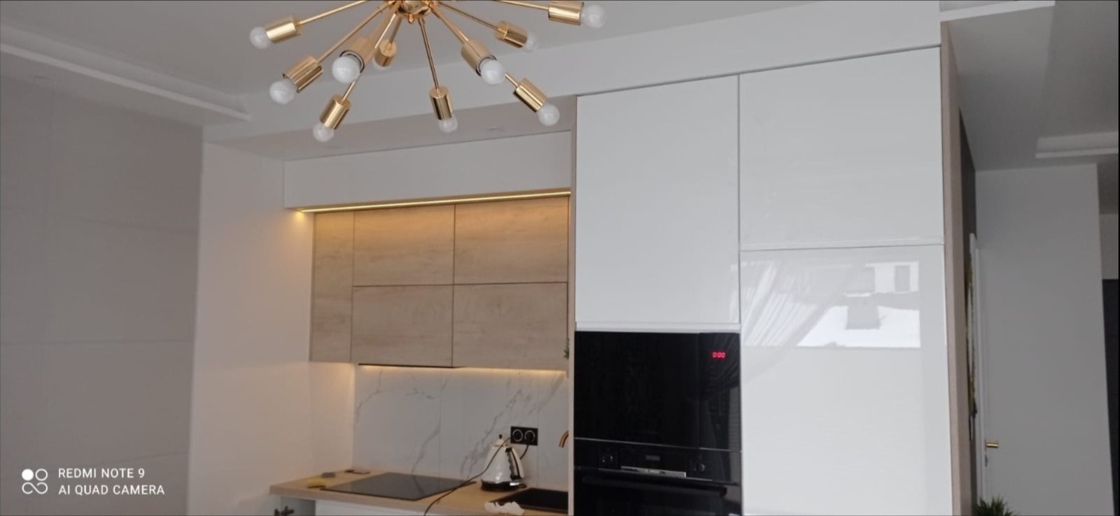 Nowoczesna kuchnia w Rzykach z białymi, lakierowanymi frontami i drewnianymi szafkami górnymi z podświetleniem LED. Marmurowy backsplash i designerska lampa.