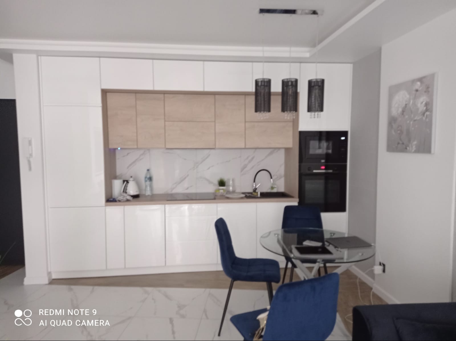 Nowoczesna kuchnia z białymi szafkami i drewnianymi akcentami, marmurowy backsplash, okrągły stół ze szklanym blatem i niebieskimi krzesłami, minimalistyczne lampy wiszące.