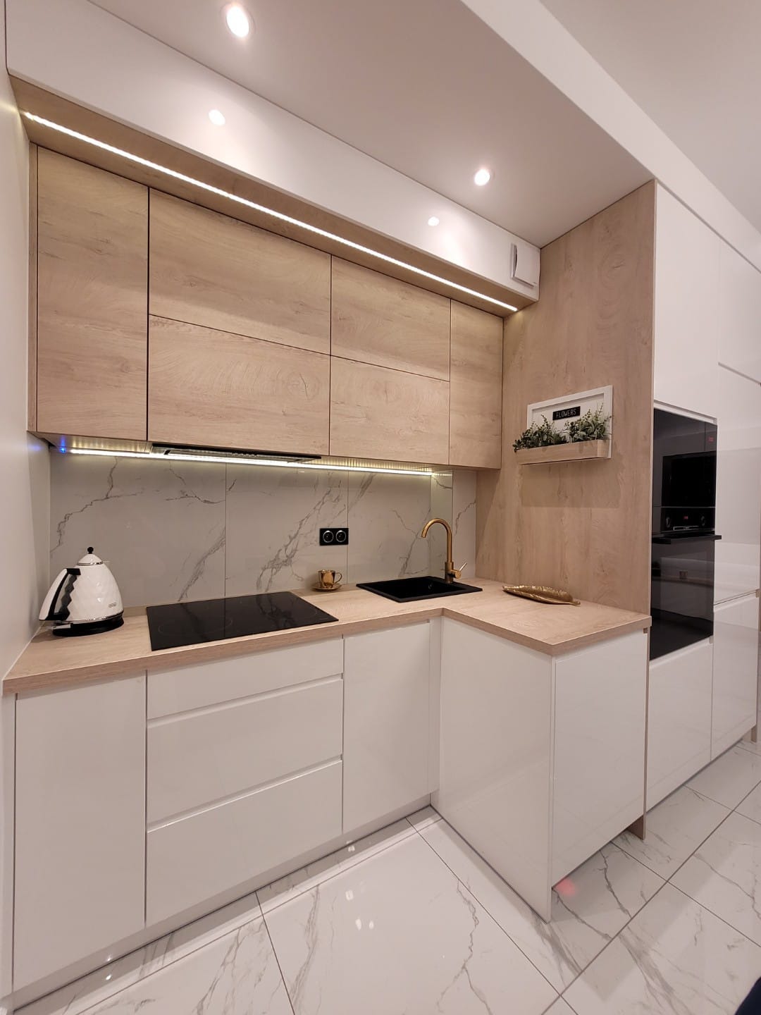 Nowoczesna kuchnia z białymi, lakierowanymi frontami i drewnianymi szafkami górnymi z oświetleniem LED. Marmurowe płytki na ścianie i podłodze.