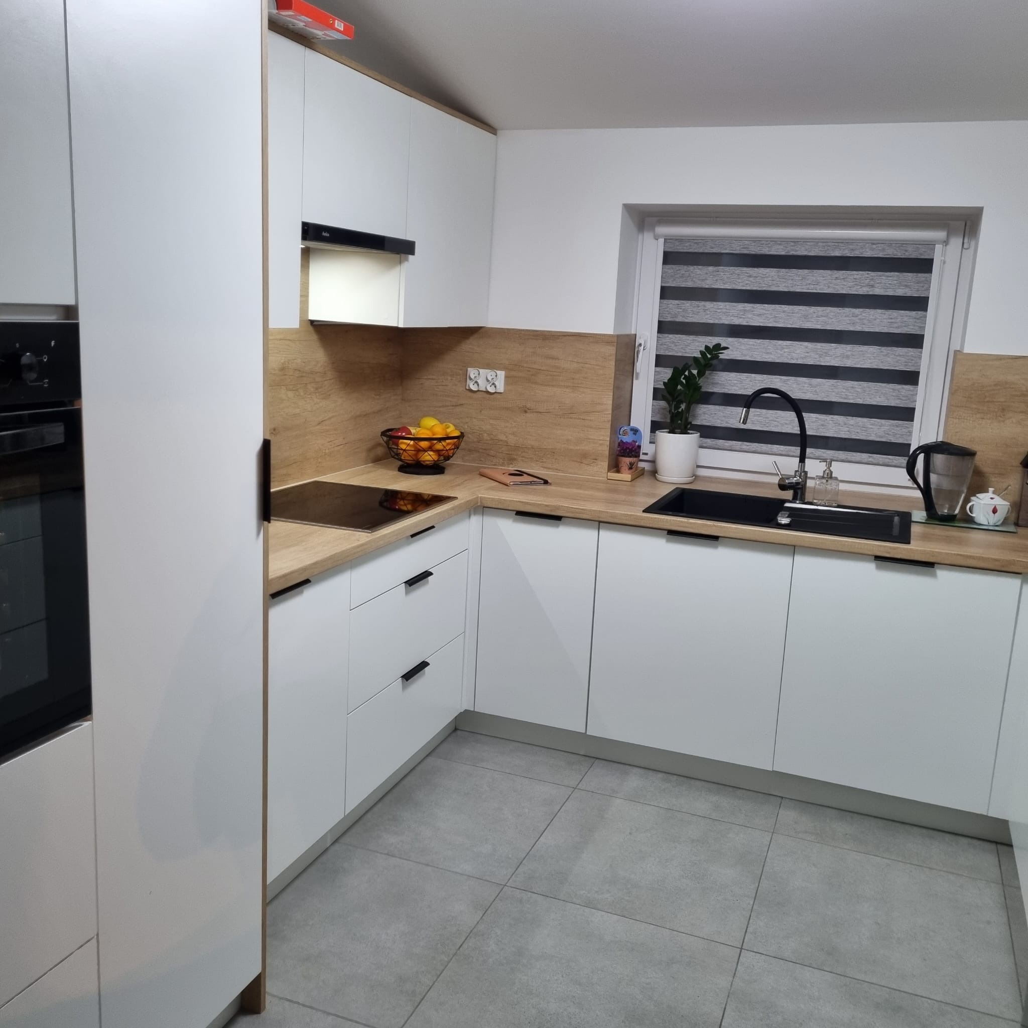 Biała kuchnia z drewnianym blatem i czarnymi uchwytami, widok na zlew, płytę indukcyjną i okno z roletami dzień-noc. Na blacie owoce i akcesoria kuchenne.