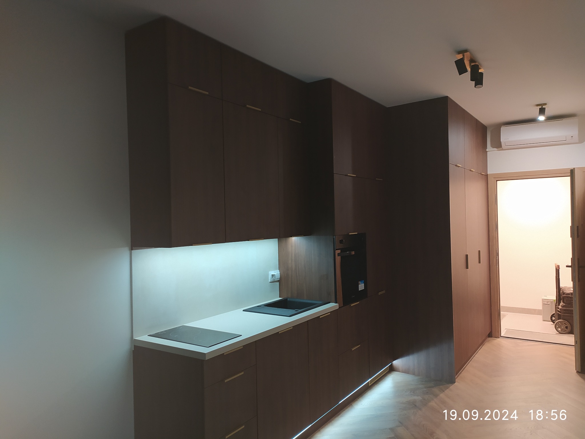 Ciemna zabudowa kuchenna z oświetleniem LED pod szafkami i blatem, widok na korytarz z podłogą w jodełkę. Minimalistyczny design z długimi, cienkimi uchwytami.