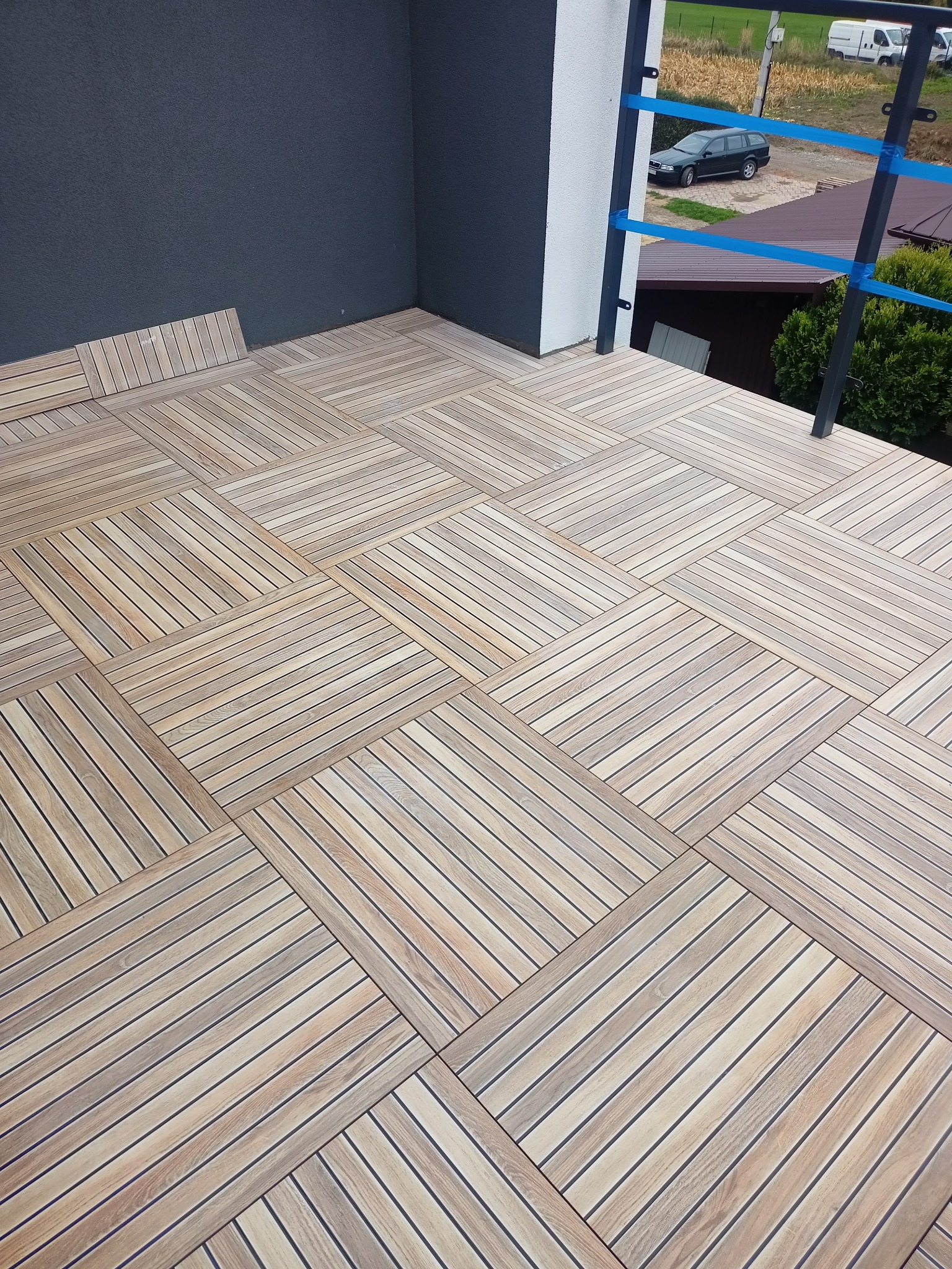 Wyłożona płytkami imitującymi drewno podłoga balkonu z widokiem na podwórze i zaparkowany samochód.